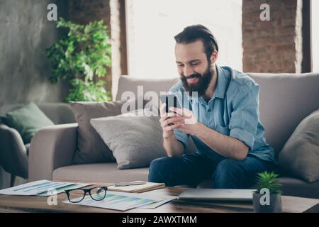 Portrait seines hübschen fröhlichen fröhlichen brunet-kerls, der auf Divan sitzt, der SMS tippt, mit Freundin im Industrial Loft modern Style chattet Stockfoto