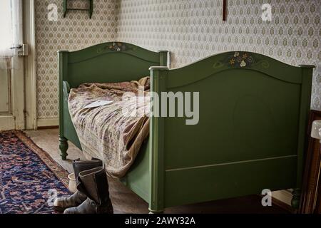 Vintage-Bett in einem Schlafzimmer mit gemusterten Tapeten und abgenutzten Stiefeln auf einem Teppich und Holzboden Stockfoto