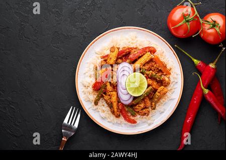 Gemüsejalfrezi mit Basmati-Reis in weißer Platte auf schwarzem Schieferhintergrund. Jalfrezi ist ein Gericht der indischen Küche mit gebratenem Gemüse und Tomaten-n Stockfoto