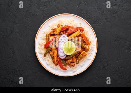 Gemüsejalfrezi mit Basmati-Reis in weißer Platte auf schwarzem Schieferhintergrund. Jalfrezi ist ein Gericht der indischen Küche mit gebratenem Gemüse und Tomaten-n Stockfoto