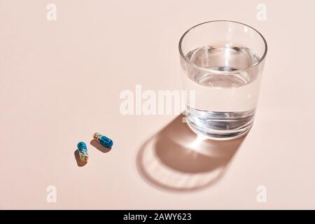 Zwei Kapseltabletten und Glas mit Wasser auf blassrosa Oberfläche, horizontaler Schuss Stockfoto