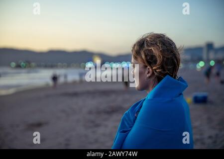 Frau beobachtet Sonnenuntergang am Strand, in blaue Decke gewickelt Stockfoto