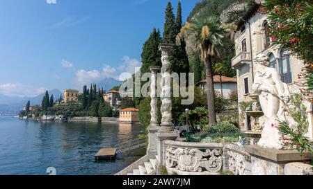 Malerische Aussicht auf die Villa Monastero mit antiken Statuen, Vordersäulen und einer Marmortreppe zum Pier, umgeben von Palmen und Thuja. Ansicht eines antiq Stockfoto