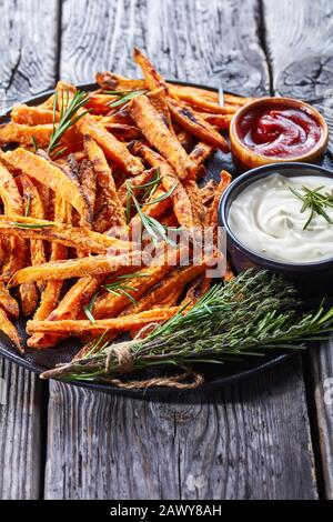 Nahaufnahme von knusprigen Süßkartoffelfries, die in einem Ofen gebacken und mit Sauce und Ketchup auf einer schwarzen Platte auf einem rustikalen Holztisch serviert werden, vertikale Ansicht Stockfoto
