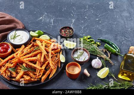 Nahaufnahme von knusprigen Süßkartoffeln mit Soße und Ketchup auf einer schwarzen Platte auf einem Betontisch mit Kalk und Zutaten, Stockfoto