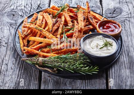 Knusprige Süßkartoffeleien, die im Ofen gebacken werden, serviert mit Sauerrahm und Ketchup auf einer schwarzen Platte auf einem rustikalen Holztisch, in der Nähe Stockfoto