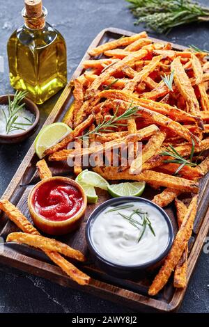 Knusprig salzig süße Kartoffelcreen mit Sauce und Ketchup auf einer unschönen Holzplatte auf einem Betontisch mit Limette und Blumenstrauß aus aromatischen Kräutern, vertikal VI Stockfoto