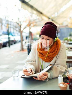 Junge Frau mit Kopfhörern, die im Herbst im Café Sidewalk im Journal schreibt Stockfoto