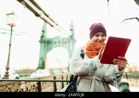 Junge Frau in Strumpfkappe und Schal mit digitalem Tablet auf der städtischen Brücke Stockfoto