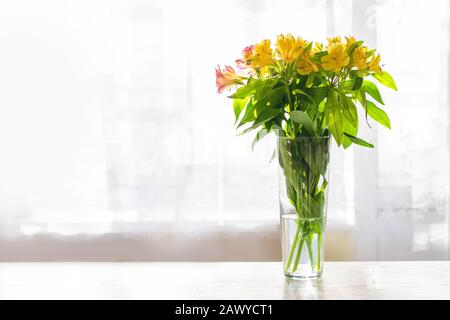 Gelbe Blumen in einer Glasvase mit Wasser auf einem weißen Tisch vor dem Hintergrund eines Fensters mit Dachpapier Stockfoto