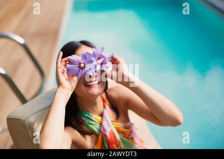 Portrait verspielte Frau, die lila Hibiskus über die Augen am sonnigen Sommer am Pool hält Stockfoto