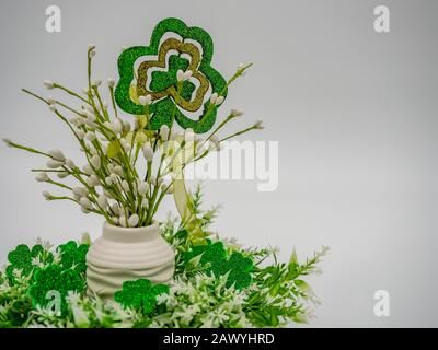 Weiße behauene Blumenarrangement umgeben von Grün und glitzernden vier Blattklüppchen mit einem großen vierblättrigen Kleeblatt und einem grünen Bogen auf einer weißen Rückengrosche Stockfoto