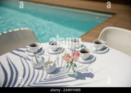 Kaffeetassen und Rosen auf dem sonnigen Terrassentisch am Pool Stockfoto