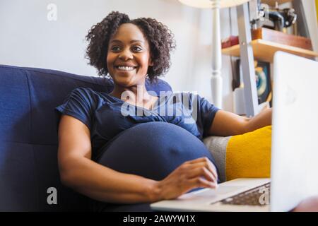 Glückliche schwangere Frau mit Laptop auf dem Sofa Stockfoto