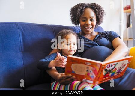 Schwangere Mutter liest Buch zur Tochter auf dem Sofa Stockfoto