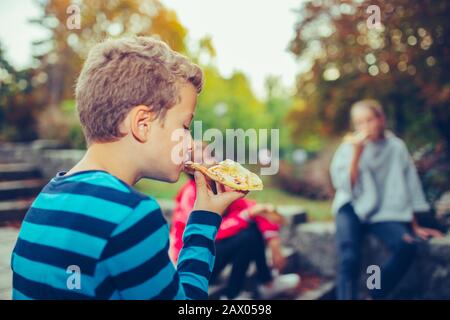 Kleiner glücklicher Junge, der im Freien Pizza isst, während er mit seinen Freunden im Park sitzt Stockfoto