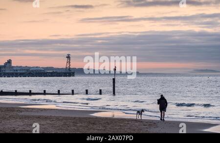 Mann und Hund machen einen frühen Morgenspaziergang im Morgengrauen am Meer in Bournemouth Stockfoto