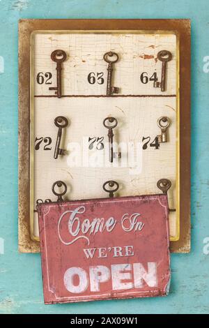 Kleines Vintage-Kabinett mit verrosteten Hotelschlüsseln und Zimmernummern und kommen herein wir sind offenes Schild auf einem blau erodierten Hintergrund Stockfoto