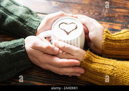 Nahaufnahme eines Paares, das die Hände an einer Tasse Kaffee mit einem Kakaoherz auf Milchschaum hält Stockfoto