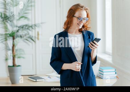 Innenschuss von positiv rothaarigen Frauen überprüft Emialbox auf modernen Smartphone-Gerät, trägt optische Brille, verwendet moderne Technologien, erhält gut Stockfoto