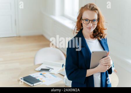 Schuss der schönen, rothaarigen jungen Frau überprüft die Benachrichtigung auf dem Touchpad, freut sich, Einkommensnachrichten zu erhalten, schaut selbstbewusst auf Kamera, Arbeitsplatz Witz Stockfoto