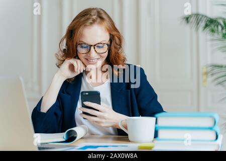 Foto einer hübschen Frau sitzt mit Smartphone-Gerät, gibt Feedback, arbeitet im Büro auf einem aktuellen Laptop, fokussiert auf den Bildschirm von Gadgets, sitzt bei der Arbeit Stockfoto