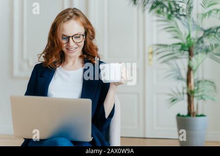 Zufriedene europäische Frauen mit Ingwerhaar, Typen von Informationen auf dem Laptop, hat einen frohen Gesichtsausdruck, trinkt Kaffee, trägt Brille, entwickelt sich Stockfoto