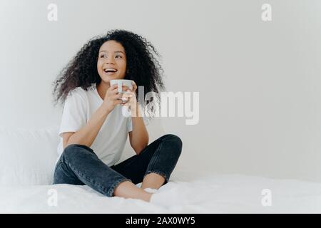 Faules Konzept für morgens und abends. Die positive afro-Frau hält einen Becher heißen Tees oder Kaffees, sitzt im Bett, sieht schön beiseite, genießt gutes Wetter, trägt c Stockfoto