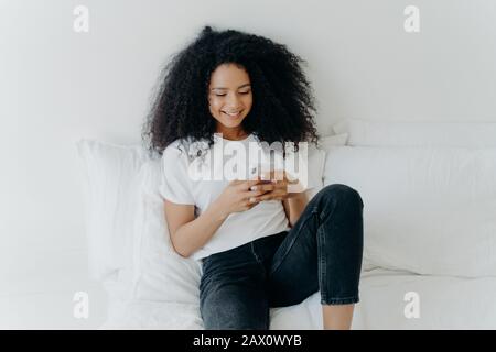 Die entspannte, schöne afro-Frau mit fröhlichem Ausdruck verwendet modernes Handy, surft Internet, posiert in weißem, bequemem Bett, verbunden mit wifi, bro Stockfoto