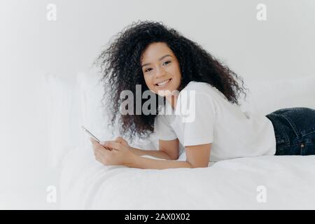Hübsche fröhliche Frau mit Afro Haarschnitt, genießt Plaudern und Networking, liegt auf bequemem Bett, sieht Fotos aus, hat entspannten Gesichtsausdruck, isoliert Stockfoto