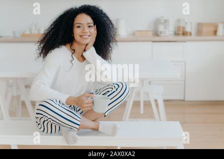 Innenschuss der hübschen afroamerikanischen Frau trägt weißen Pullover, gestreifte Hosen, Socken, Posen auf der Bank mit einer Tasse Tee verbringt Freizeit zu Hause in gemütlichem k Stockfoto