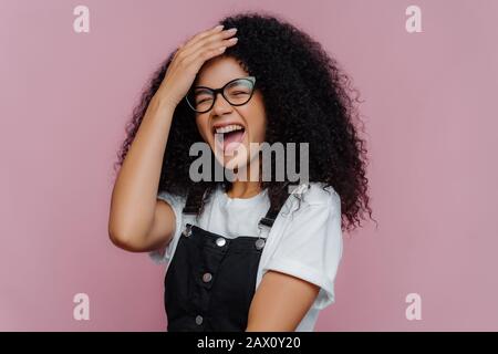 Überfreudig geflechte, lockig behaarte Frau hält Hand auf Stirn, lacht vom Glück, versteht, dass sie etwas komisch gemacht hat, trägt optische Brille, lässig Stockfoto