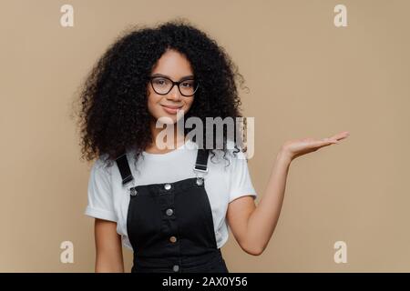 Horizontaler Schuss von ziemlich dunkelhäutigem geschweiften Mädchen hebt Handfläche an, hält freien Platz, trägt transparente Brille, weißes T-Shirt und Overalls, isoliert davon Stockfoto