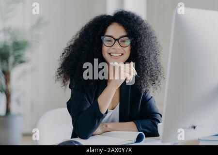 Positive internationale Studentin mit knackigen Haaren, trägt transparente Brille, hält Stift in der Hand, macht Buchhaltung, sitzt vor dem großen Computer Stockfoto