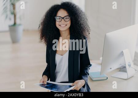 Gefiel finstere, dunkelhäutige geschweifte Frau hält einige Papiere, Posen im modernen Büroinneren, trägt transparente Brille und formellen Anzug, steht in der Nähe von TA Stockfoto