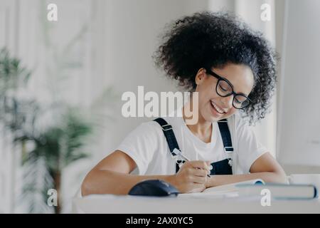 Die fröhliche afro-amerikanische Studentin schreibt Informationen auf, bereitet sich auf Prüfungen vor, sitzt im feigen Raum, hat geschweifte dunkle Haare, trägt eine optische Brille WH Stockfoto