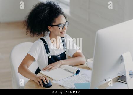 Die junge afroamerikanische Frau, die froh ist, sitzt vor dem modernen Computer, macht Projektarbeit, hat viele Papiere und Notizblöcke auf dem Tisch, trägt Brille für Vis Stockfoto