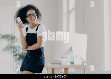 Fröhliches, lockig behaartes Mädchen verwendet Handy für Kommunikation und Kontakt, trägt Brille und legere Kleidung, lehnt sich am Schreibtisch, posiert in Spac Stockfoto