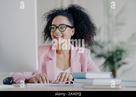 Foto der froh geschweiften professionellen weiblichen Managerin, die sich auf den Computerbildschirm konzentriert, lächelt breit, hat lockiges Haar, trägt transparente Brille, eleganten Anzug, Stockfoto