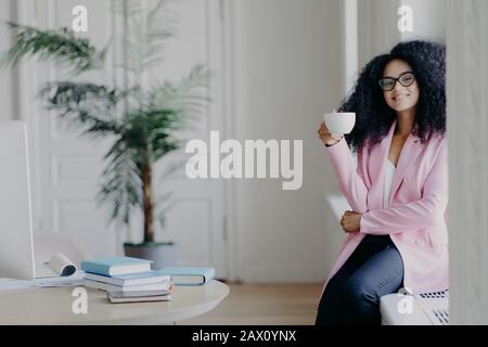 Die positive afroamerikanische Arbeitskraft für Frauen sitzt auf der Fensterbank, hält einen Becher Kaffee, gekleidet in formeller Kleidung, stellt sich in der Nähe von Desktop-PCs mit Büchern, gr Stockfoto