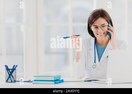 Der froh-professionelle Arzt, der sich auf den modernen Laptop konzentriert, nützliche Informationen liest, telefonische Gespräche führt, medizinische Probleme diskutiert, sitzt Stockfoto