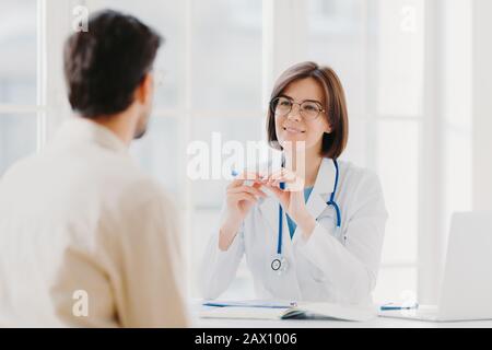 Professionelle weibliche Ärztin im Mantel hört aufmerksam die Symptome der Patienten ab, untersucht die Gesundheit, schreibt Informationen in der Patientenkarte auf, posiert in der Klinik. Si Stockfoto