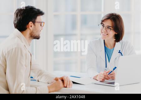 Die erfahrene professionelle weibliche Allgemeinmedizinerin erklärt die Diagnose und berät den Patienten, zeigt Informationen in Laptops, posiert im klinischen Büro Stockfoto