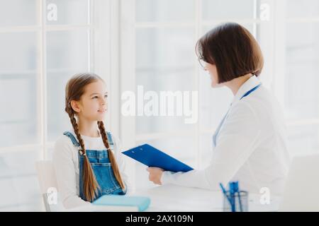 Die Kinderärztin untersucht kleine Kinder, hört sich kleine Kinder sorgfältig an, schreibt Notizen in der Zwischenablage, posieren in der Klinik oder im Krankenhaus dagegen Stockfoto