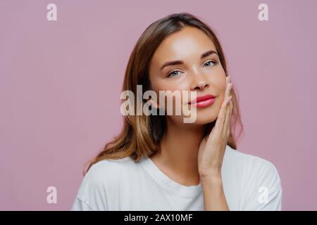 Der Kopfschuss des hübschen weiblichen Models berührt sanft Wange, genießt zarte Gesichtshaut, trägt Make-up, hat Maniküre, kümmert sich um ihre Schönheit, sieht gerade aus Stockfoto