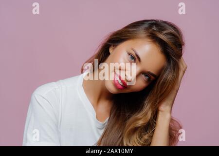 Portrait des gut aussehenden, freudigen weiblichen Models neigt den Kopf, hat ein zottiges Lächeln zeigt weiße perfekte Zähne, trägt einen legeren Pullover, berührt Haare, zeigt ihr bea Stockfoto