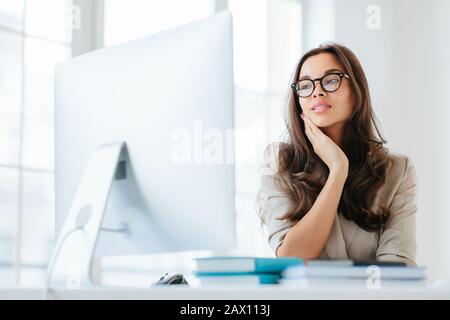 Innenaufnahme einer ernsthaften, auf Computerüberwachung konzentrierten Brunette-Dame, die zur Veröffentlichung drahtloses Internet verwendet, die Hand auf Wange hält, einen aufmerksamen Blick hat, Stockfoto