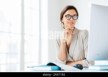 Die charmante Büroangestellte hält die Hand unter dem Kinn, schaut auf dem Bildschirm des Computers, trägt Brille zur Korrektur der Sehkraft, stellt sich im eigenen Schrank vor, bereitet vor Stockfoto