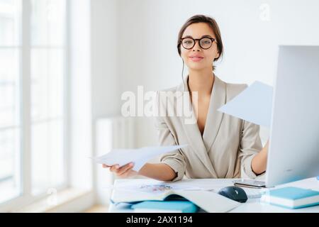 Viel erfolgreiche Managerin arbeitet mit Papieren im Büro, posiert am Desktop, trägt Brille und formales Outfit, beschäftigt sich mit der Vorbereitung des Berichts, sieht verwirrt aus Stockfoto