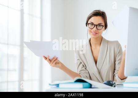 Innenaufnahmen von gut aussehenden Frauen im formalen Anzug, hält Papierdokumente, arbeitet an der Organisation und erstellt Startprojekt, stellt sich auf dem Desktop zusammen Stockfoto